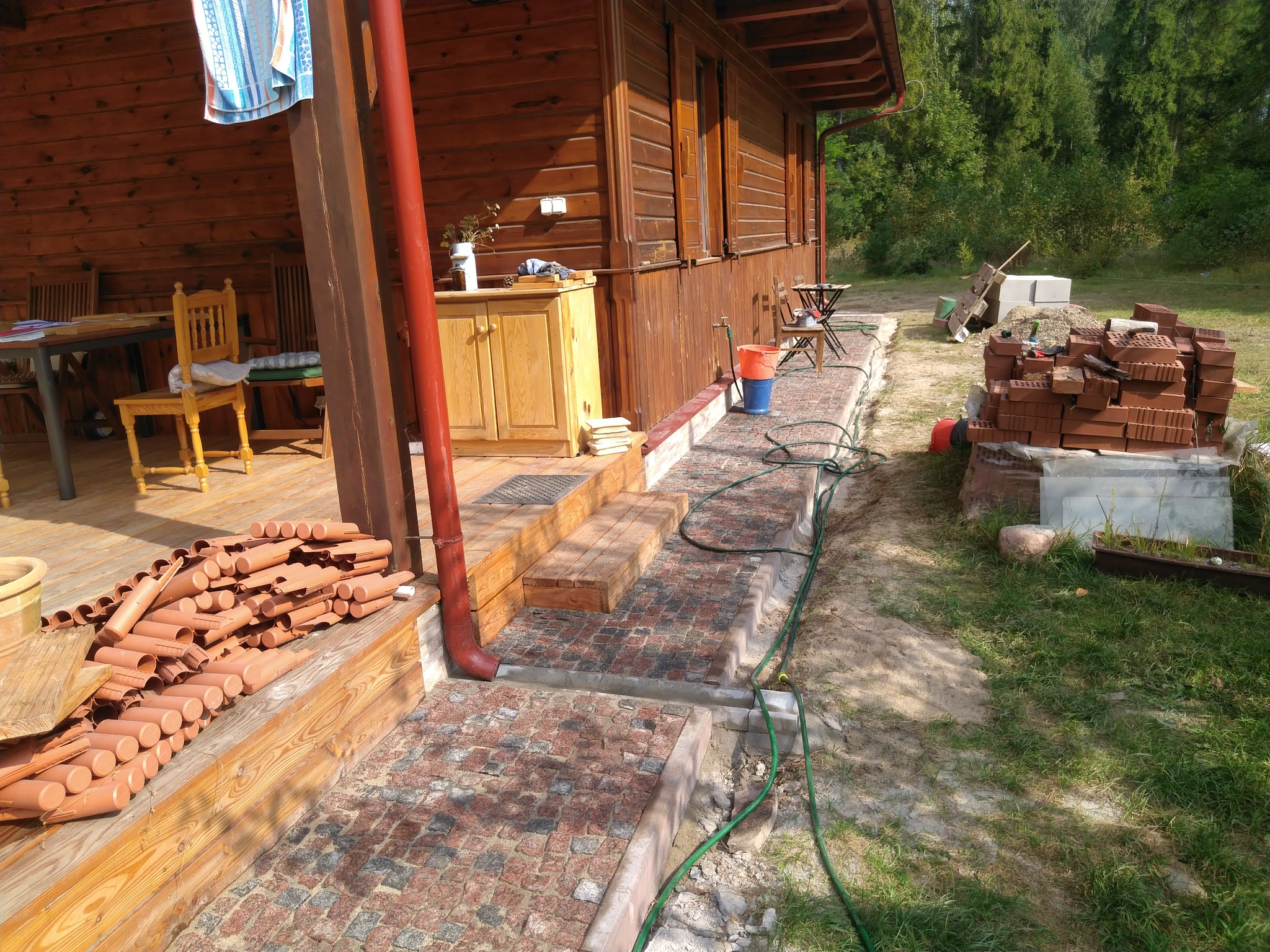 Układanie kostki brukowej z granitu na ścieżce przy drewnianym domu, widoczne przygotowane rury drenarskie i narzędzia.