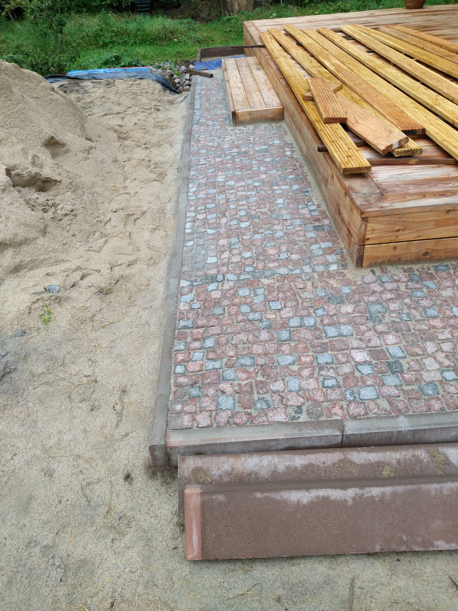 Budowa tarasu z drewnianą konstrukcją i kostką granitową, widoczny piasek przygotowany do prac ziemnych.