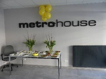 Stół z przekąskami, bukietami kwiatów i żółtymi balonami przed ścianą z napisem 'metrohouse' w nowoczesnym biurze.