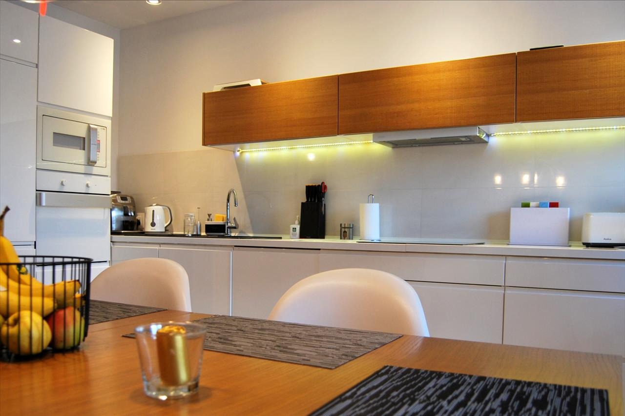 Nowoczesna, jasna kuchnia z drewnianymi szafkami górnymi podświetlonymi LED, białe fronty szafek dolnych, stół z owocami i dekoracją, widok z perspektywy siedzącego przy stole.