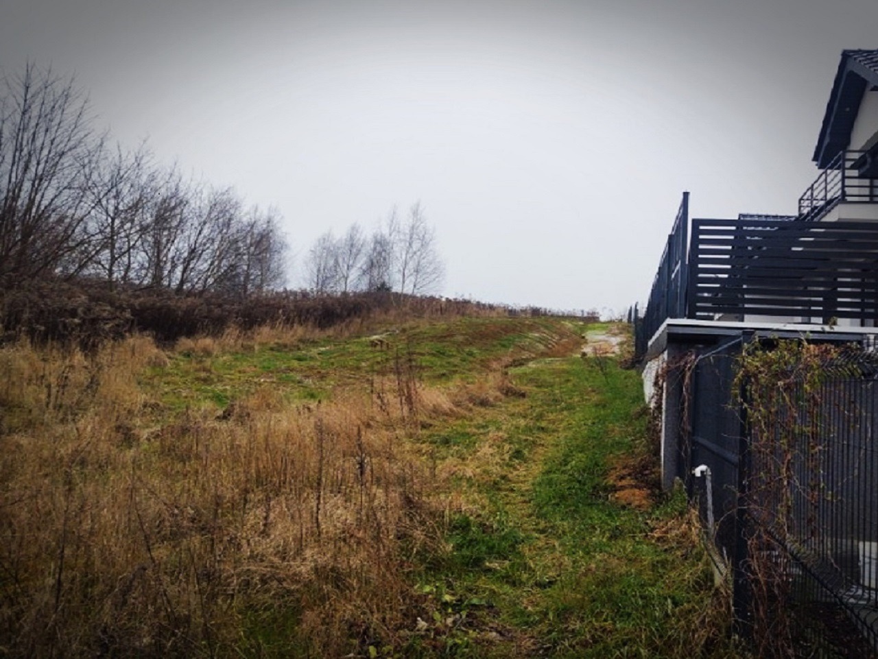 Działka budowlana z widocznymi śladami erozji zbocza, ogrodzona nowoczesnym płotem panelowym, w tle dom jednorodzinny z balkonem.