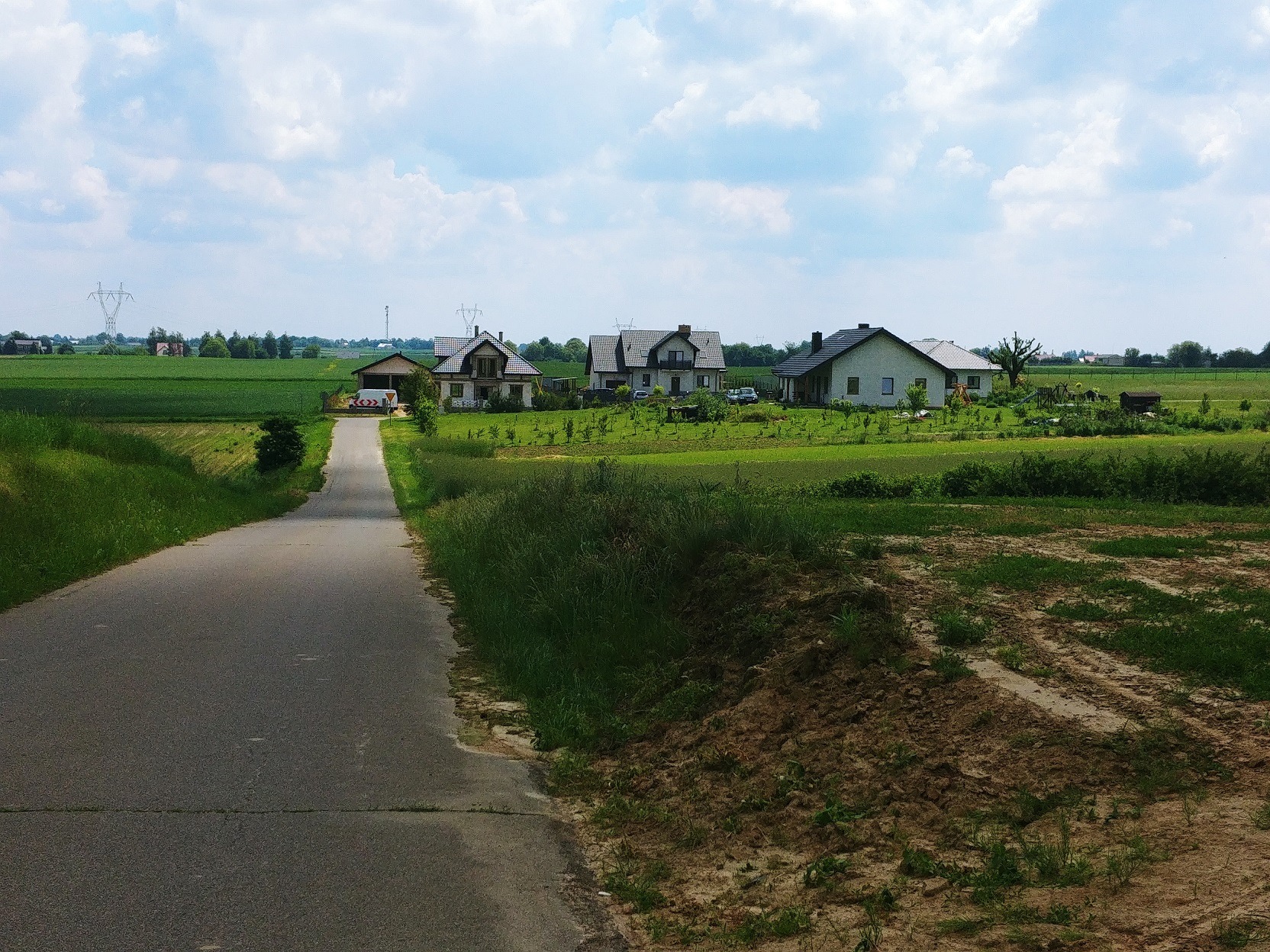 Wiejska droga prowadząca do kilku domów jednorodzinnych otoczonych polami uprawnymi i zielenią, widok w słoneczny dzień.