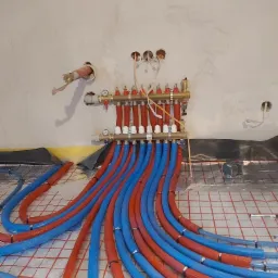 Rozdzielacz ogrzewania podłogowego z podłączonymi rurami w kolorze niebieskim i czerwonym, instalacja na siatce grzewczej, widoczne przygotowane otwory w ścianie na przewody elektryczne.