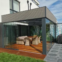 Nowoczesna szklana altana z drewnianą podłogą i meblami z rattanu, przylegająca do białego budynku z szarymi żaluzjami, otoczona trawnikiem i szarym brukiem.