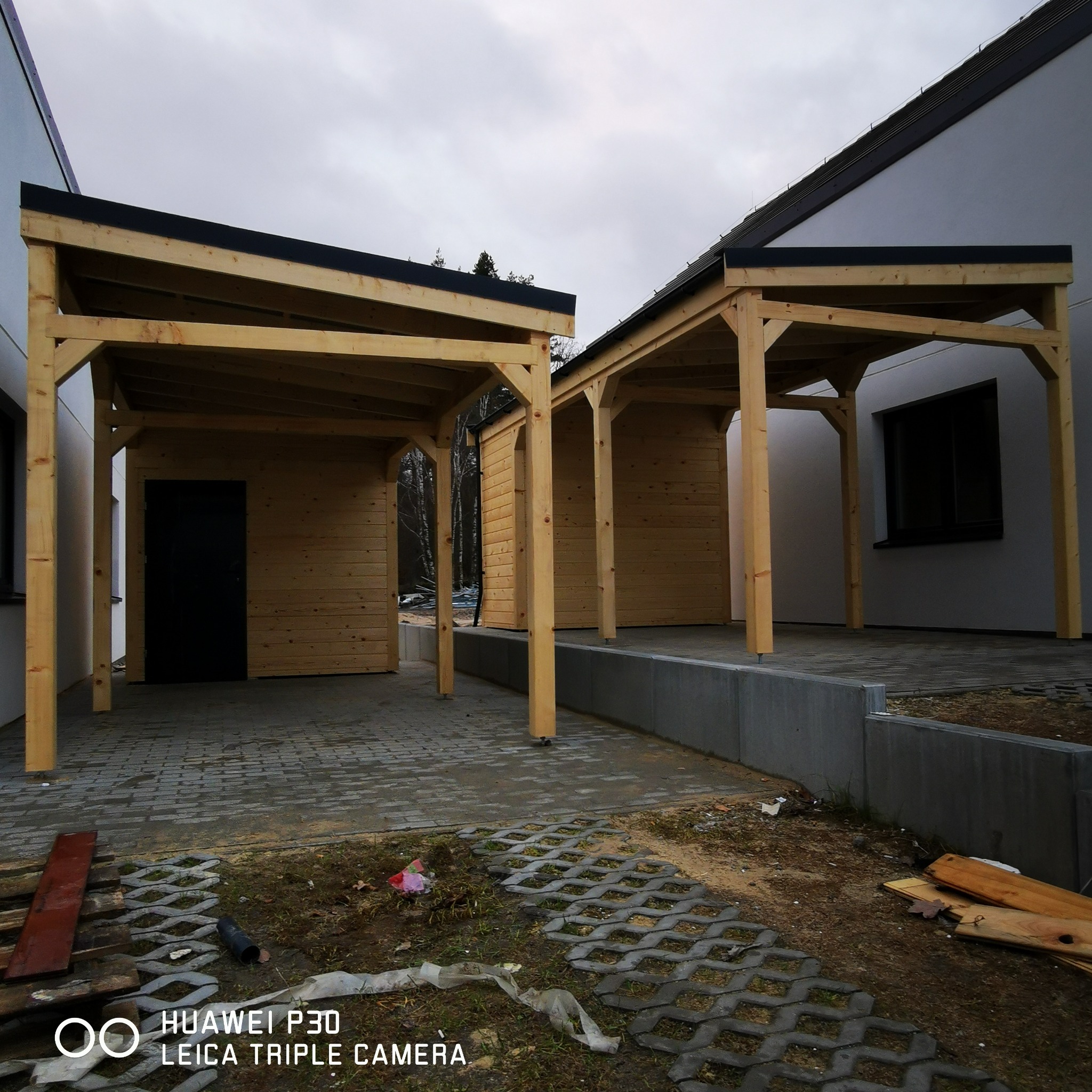 Dwa połączone drewniane carporty z czarnymi płaskimi dachami, przylegające do białych ścian budynków, widoczne z zewnątrz na tle pochmurnego nieba.