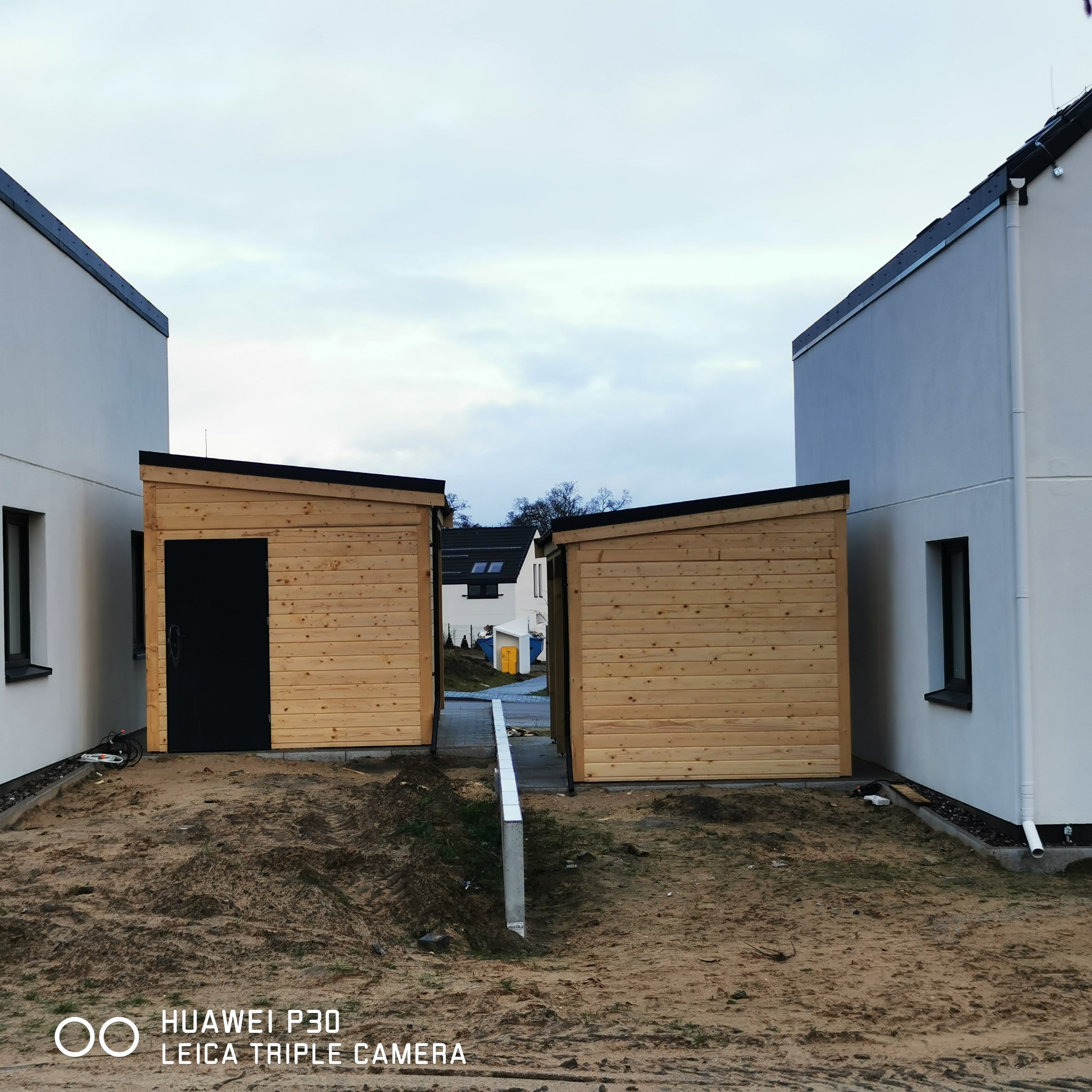 Dwa drewniane domki narzędziowe z czarnymi drzwiami, ustawione pomiędzy dwoma białymi domami jednorodzinnymi, widok z perspektywy gruntu na niezagospodarowany teren.