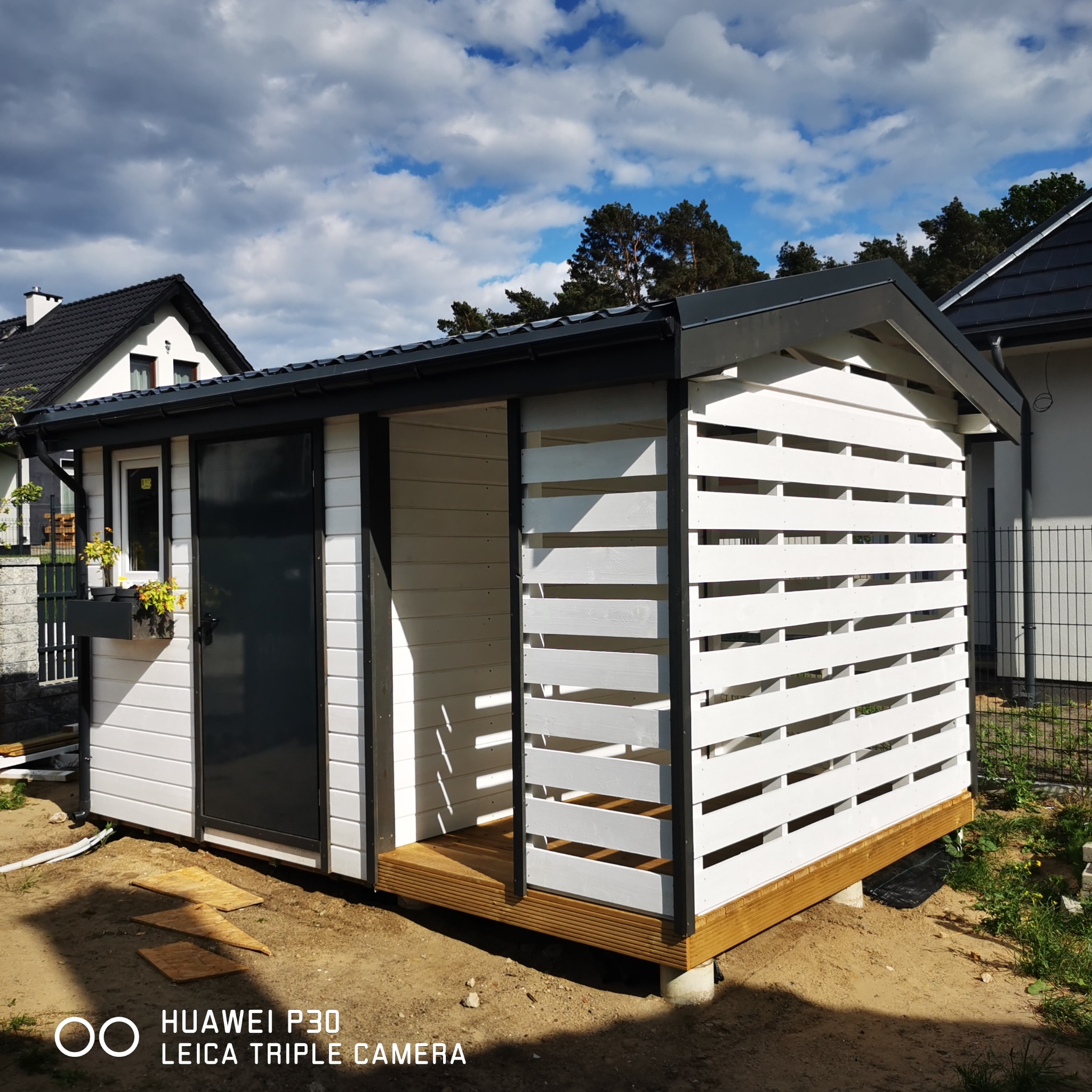 Nowoczesna, biało-czarna konstrukcja z drewnianymi lamelami, prawdopodobnie domek narzędziowy lub letniskowy, z czarnymi drzwiami i oknem z kwiatami, na betonowych fundamentach, z widocznymi domami...