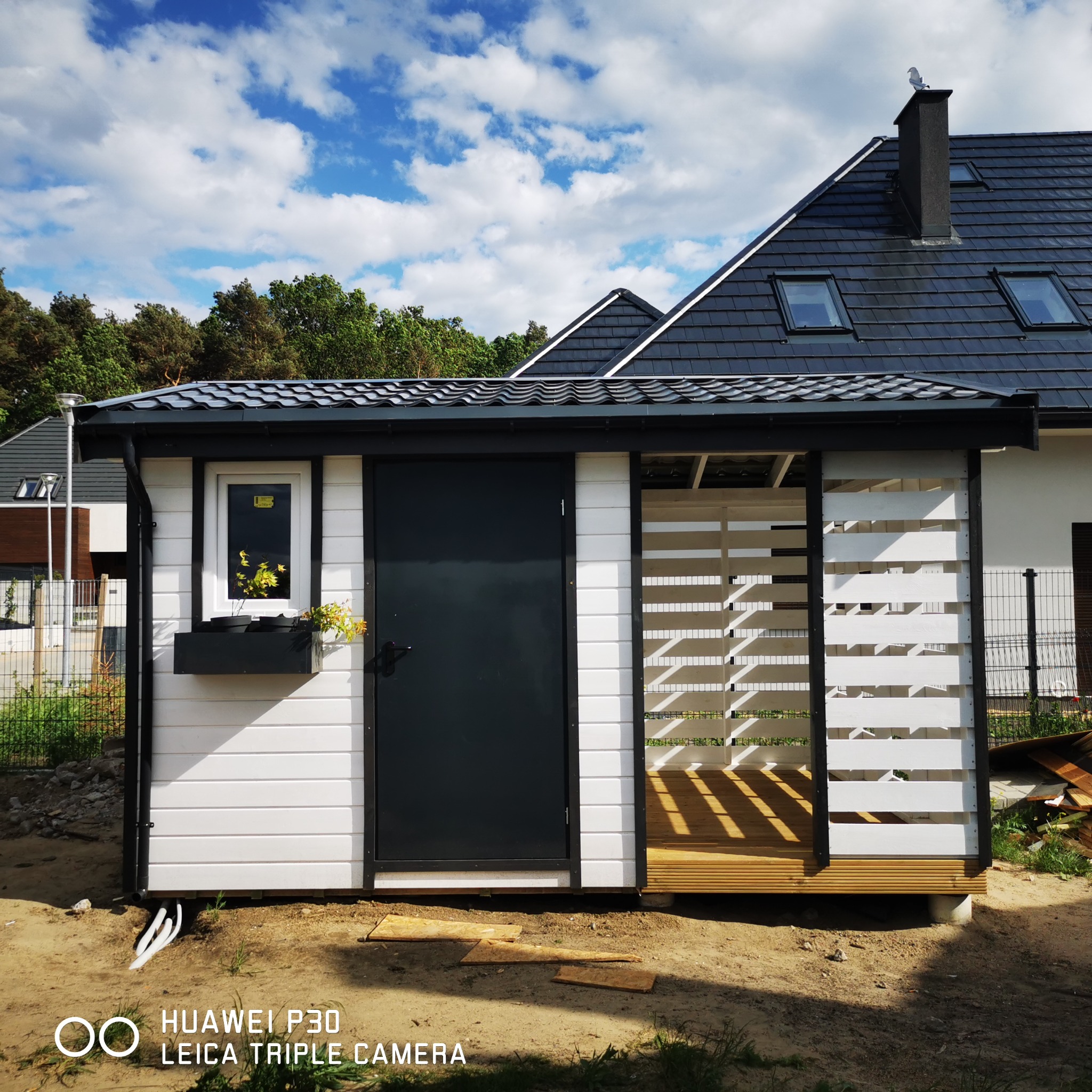 Nowoczesna, biało-czarna altana z drewnianym podestem i kratownicą, z kwiatami w oknie, na tle domu z ciemnym dachem.