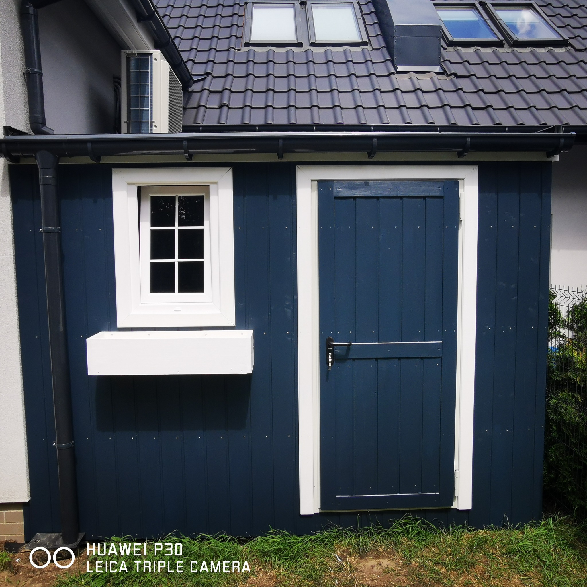 Niebieska drewniana altanka z białymi ramami okiennymi i drzwiami przesuwnymi, widoczna z zewnątrz, z oknami dachowymi na dachu domu w tle.