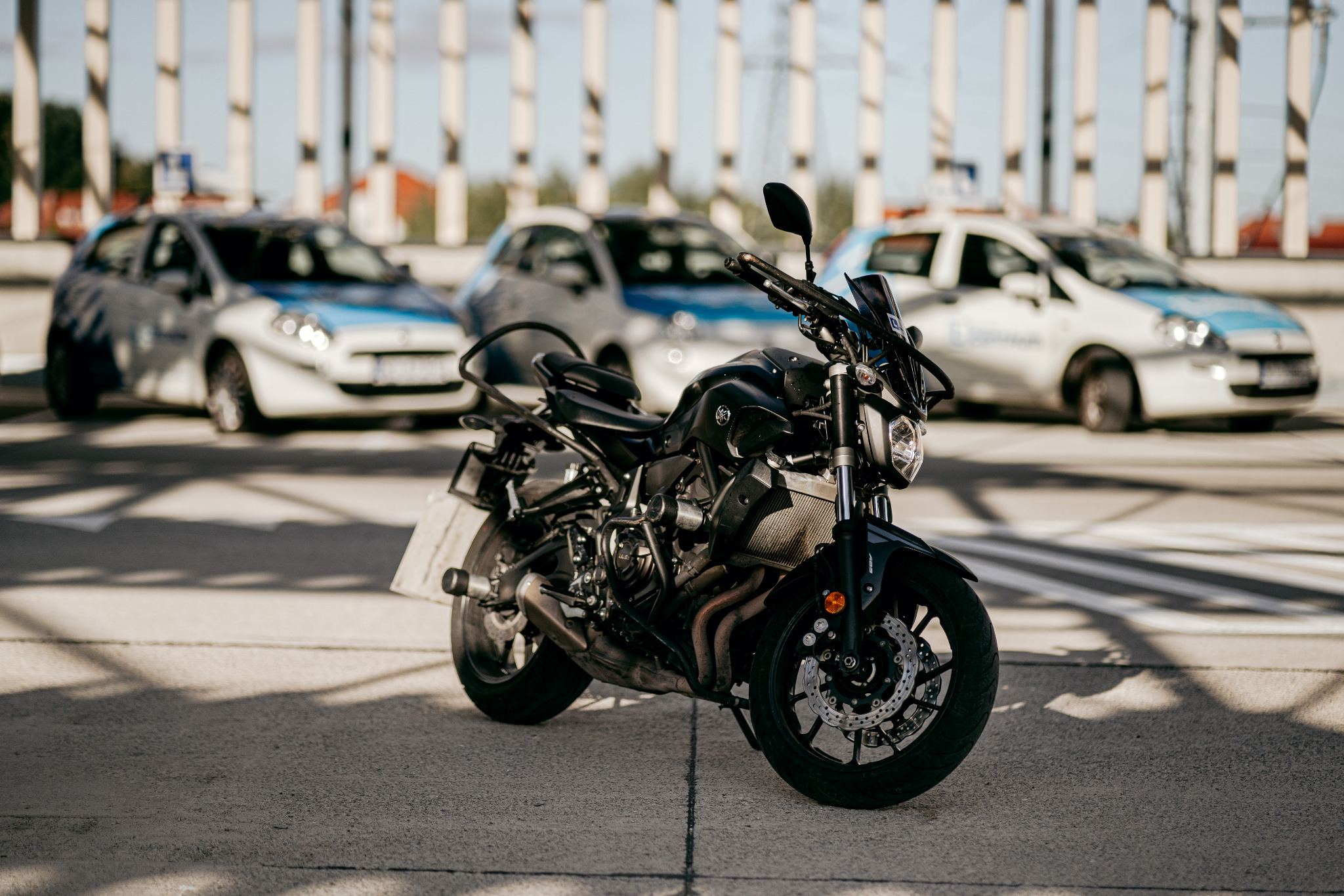 Czarny motocykl Yamaha MT-07 na tle zaparkowanych samochodów z oznaczeniami ośrodka szkolenia kierowców.