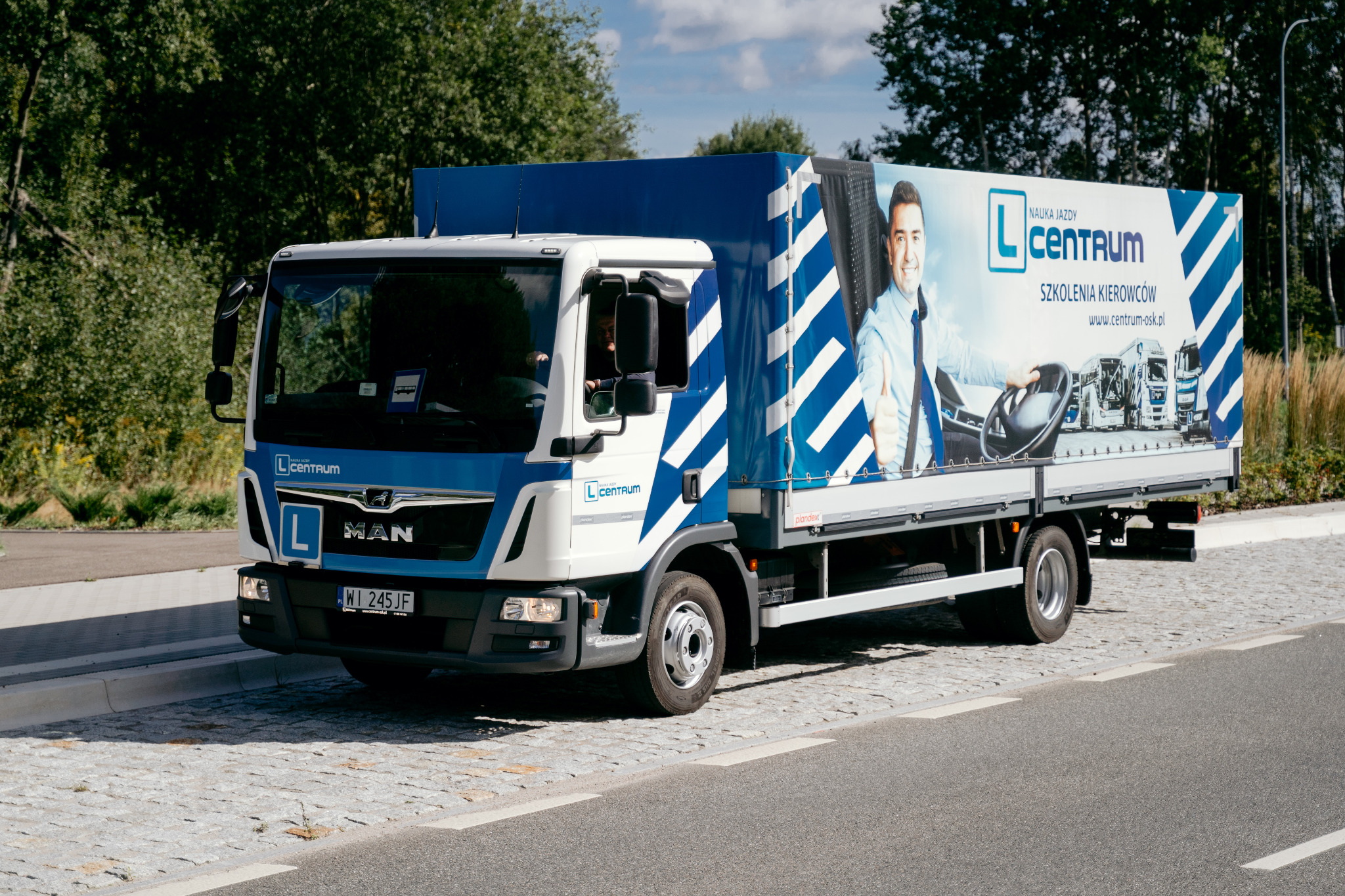 Ciężarówka MAN z niebiesko-białym brandingiem firmy szkoleniowej, z grafiką instruktora i floty na plandece, zaparkowana na kamiennej drodze.