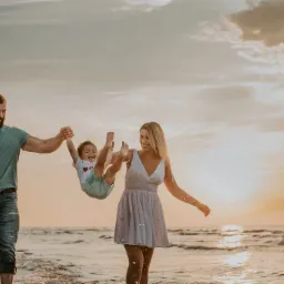 Rodzina z dwójką dzieci bawi się na plaży o zachodzie słońca; ojciec trzyma na rękach córeczkę, matka podrzuca synka.