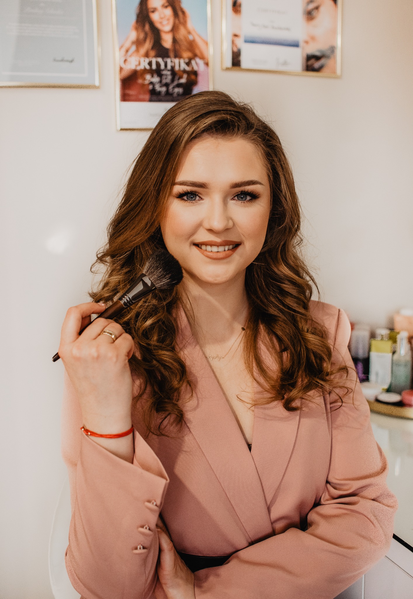 Uśmiechnięta kobieta z brązowymi włosami w różowym żakiecie trzyma pędzel do makijażu, w tle certyfikaty i kosmetyki.