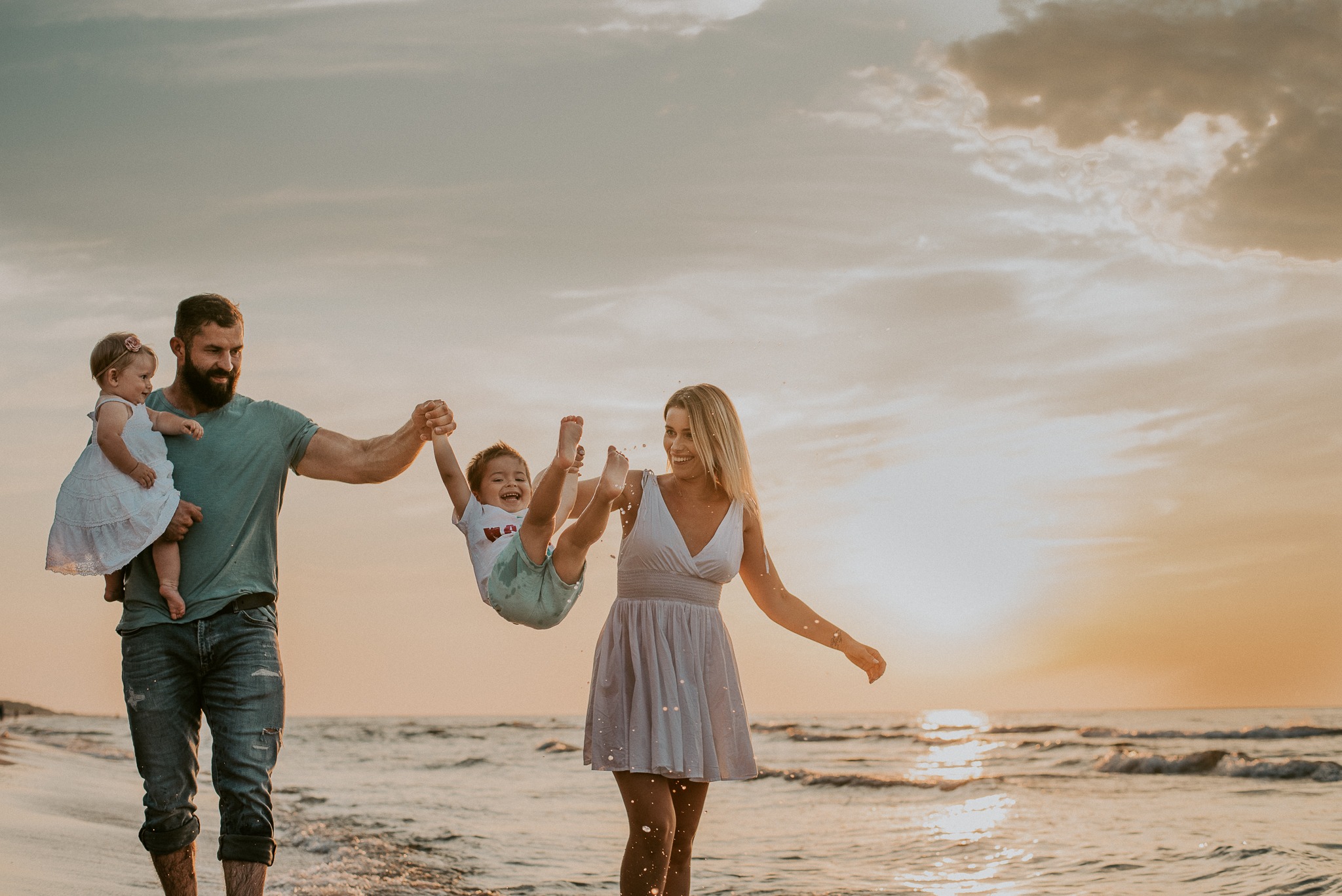 Rodzina z dwójką dzieci bawi się na plaży o zachodzie słońca; ojciec trzyma na rękach córeczkę, matka podrzuca synka.