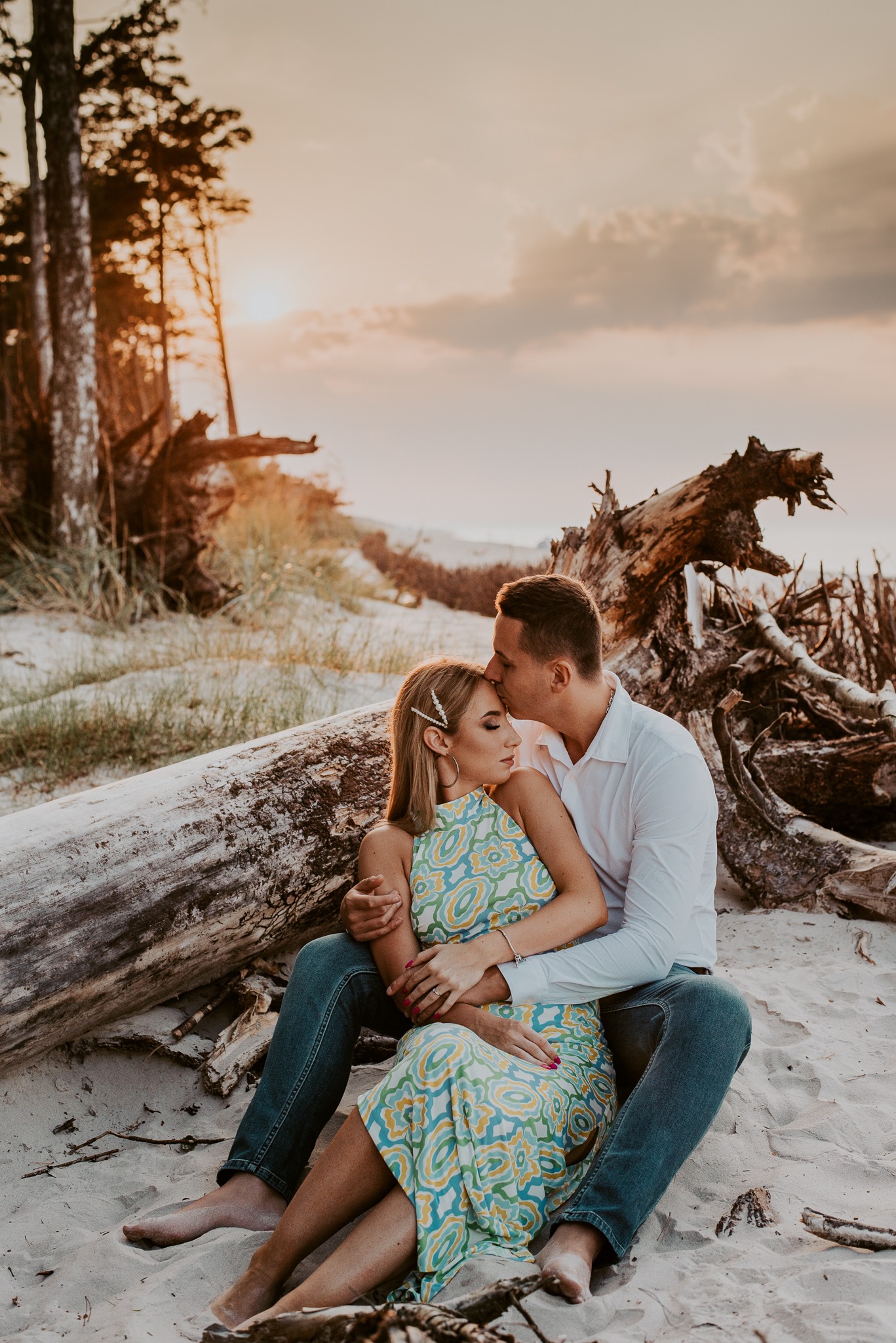 Para zakochanych siedzi na plaży przy powalonym pniu drzewa podczas zachodu słońca, mężczyzna całuje kobietę w czoło, ona ma spinkę z perełkami we włosach i sukienkę w stylu retro.