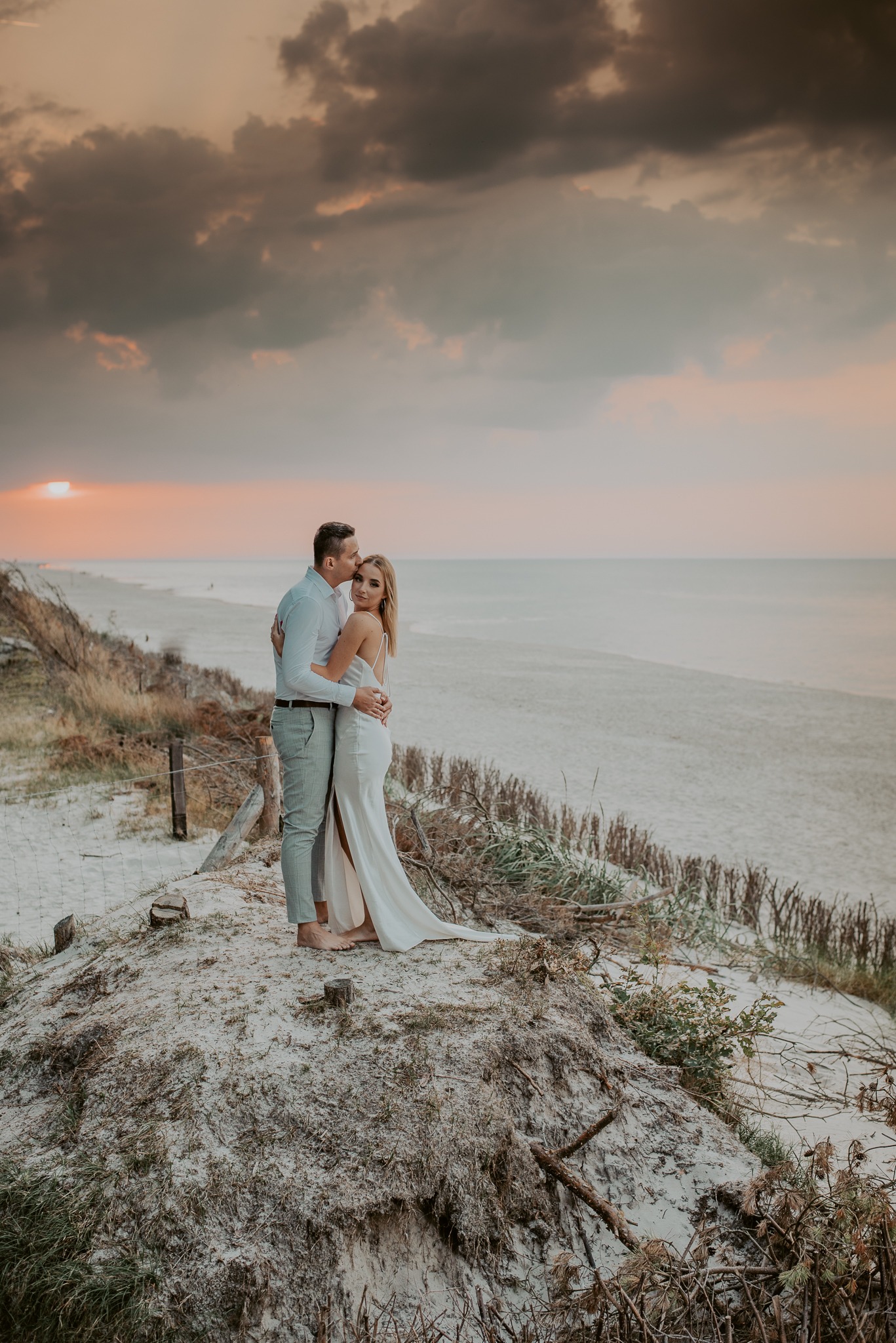 Para narzeczonych na piaszczystej wydmie przy morzu o zachodzie słońca, mężczyzna całuje kobietę w czoło, ona w białej sukni z rozcięciem.