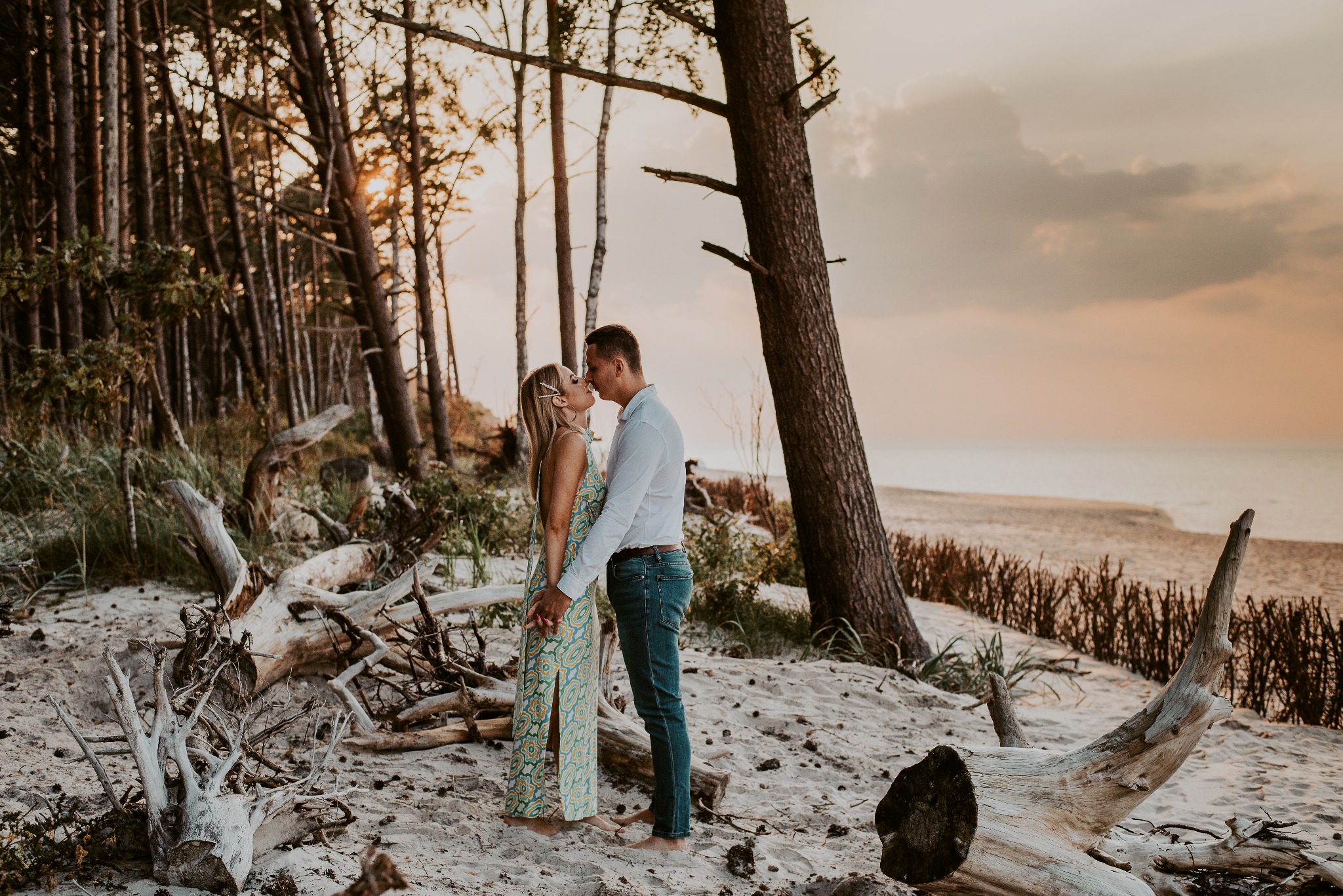 Para całująca się na piaszczystej plaży o zachodzie słońca, otoczona drzewami i leżącymi pniami, kobieta w długiej, wzorzystej sukience, mężczyzna w białej koszuli i dżinsach.