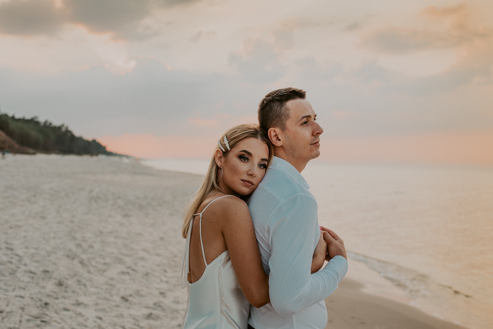 Para obejmująca się na plaży o zmierzchu; kobieta w satynowej sukience z ozdobami we włosach, mężczyzna w jasnej koszuli, oboje patrzą w dal.