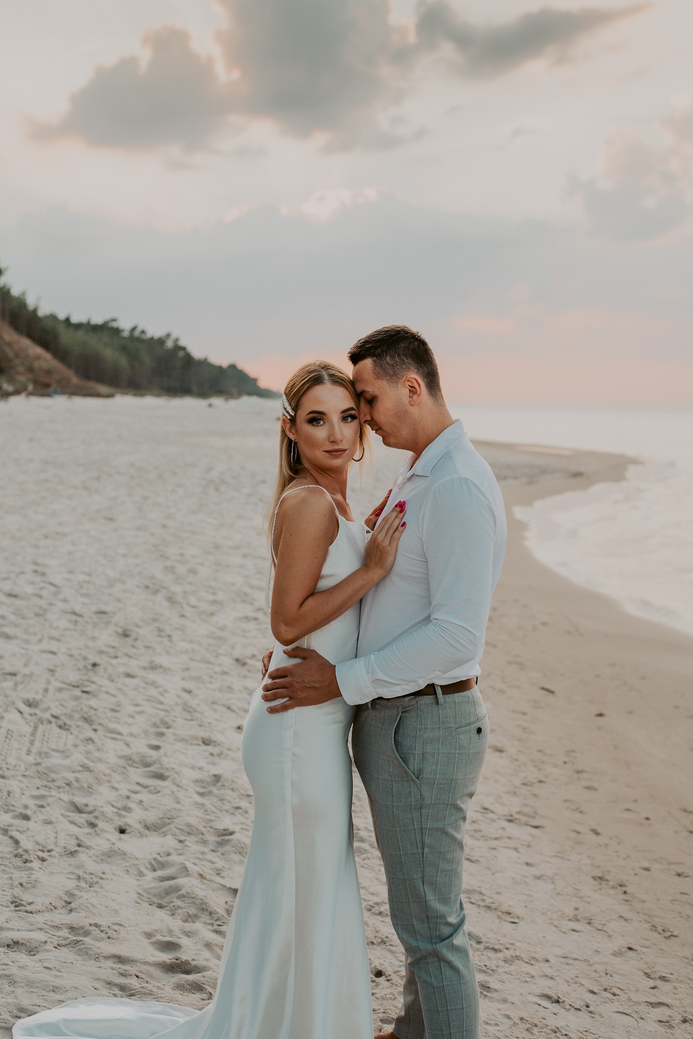 Para zakochanych na plaży o zachodzie słońca: kobieta w białej sukience z rozcięciem i ozdobą we włosach, mężczyzna w białej koszuli i szarych spodniach w kratę, delikatny uścisk.