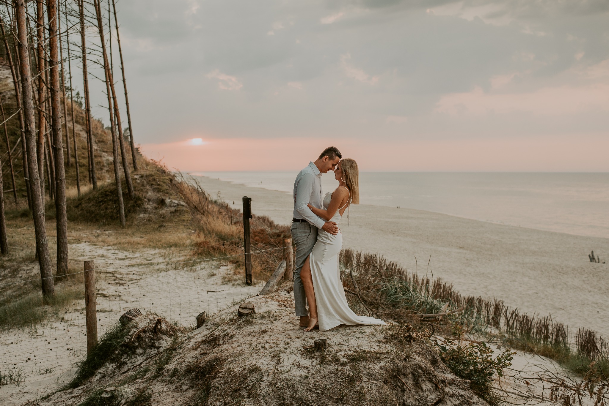 Para młoda w objęciach na tle piaszczystej wydmy i zachodzącego słońca nad morzem; on w jasnej koszuli i szarych spodniach, ona w białej sukni z odkrytymi plecami i złotych kolczykach kołach.