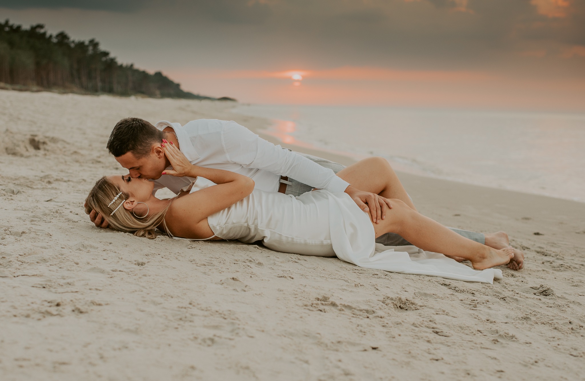 Para leżąca na piasku, całująca się na tle zachodzącego słońca nad morzem. Kobieta w białej sukience, mężczyzna w białej koszuli. Włosy kobiety upięte spinkami z perełkami.