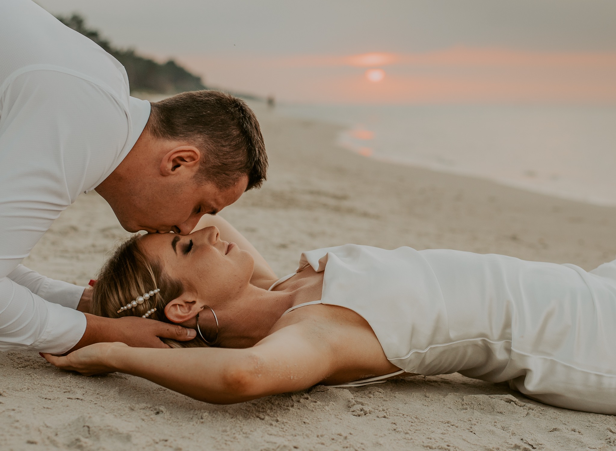 Para na plaży o zachodzie słońca: mężczyzna całuje leżącą kobietę w czoło, oboje ubrani na biało, ona z perłowymi spinkami we włosach i dużymi kolczykami kołami.