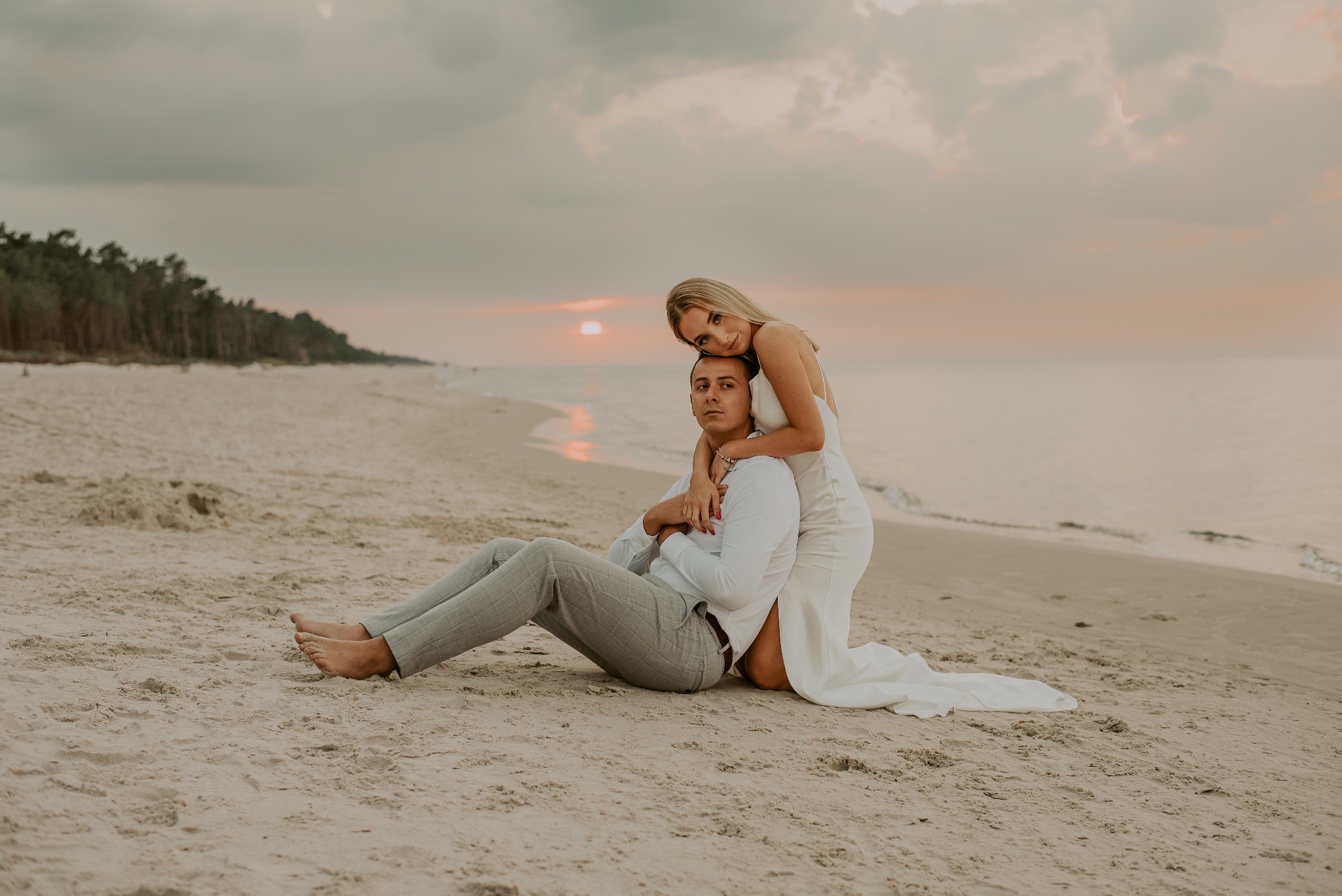 Para narzeczonych siedzi na piaszczystej plaży o zachodzie słońca, kobieta obejmuje mężczyznę od tyłu, oboje ubrani na biało z elementami szarości.
