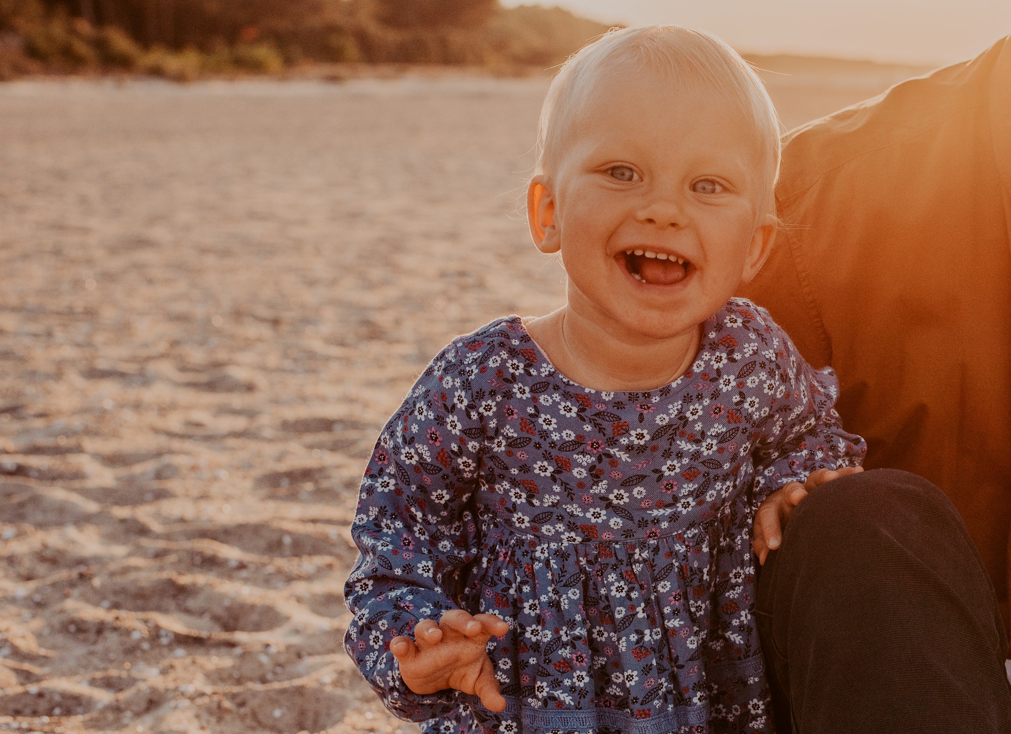Uśmiechnięta blond dziewczynka w kwiecistej sukience siedzi na plaży obok osoby dorosłej, ciepłe światło zachodzącego słońca.