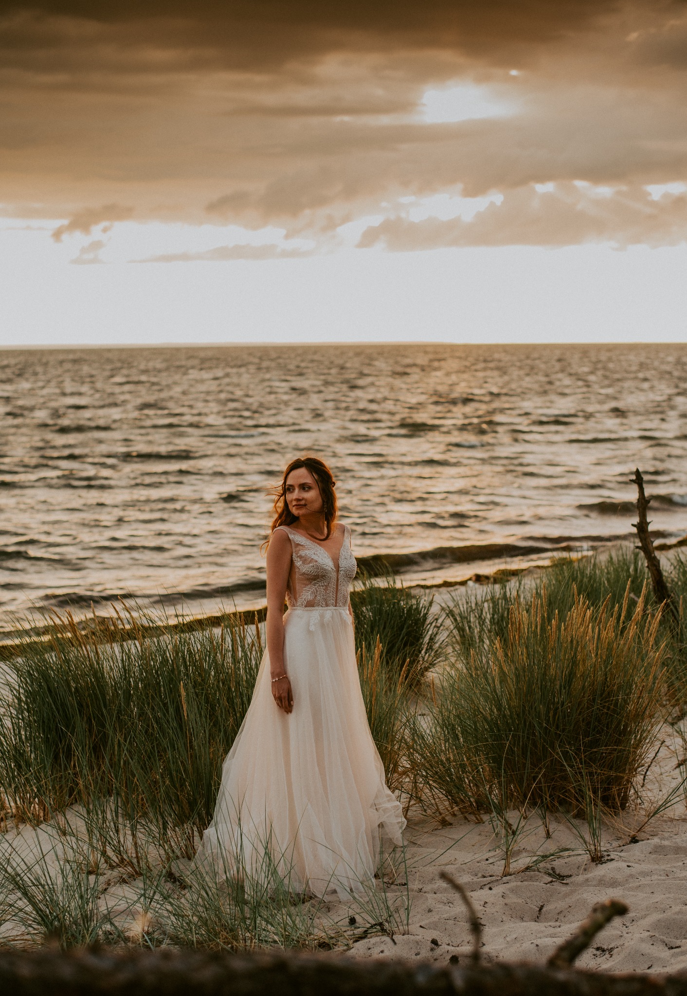 Panna młoda w zwiewnej sukni ślubnej z koronkową górą, pozująca na piaszczystej plaży wśród traw, z widokiem na morze i zachodzące słońce.