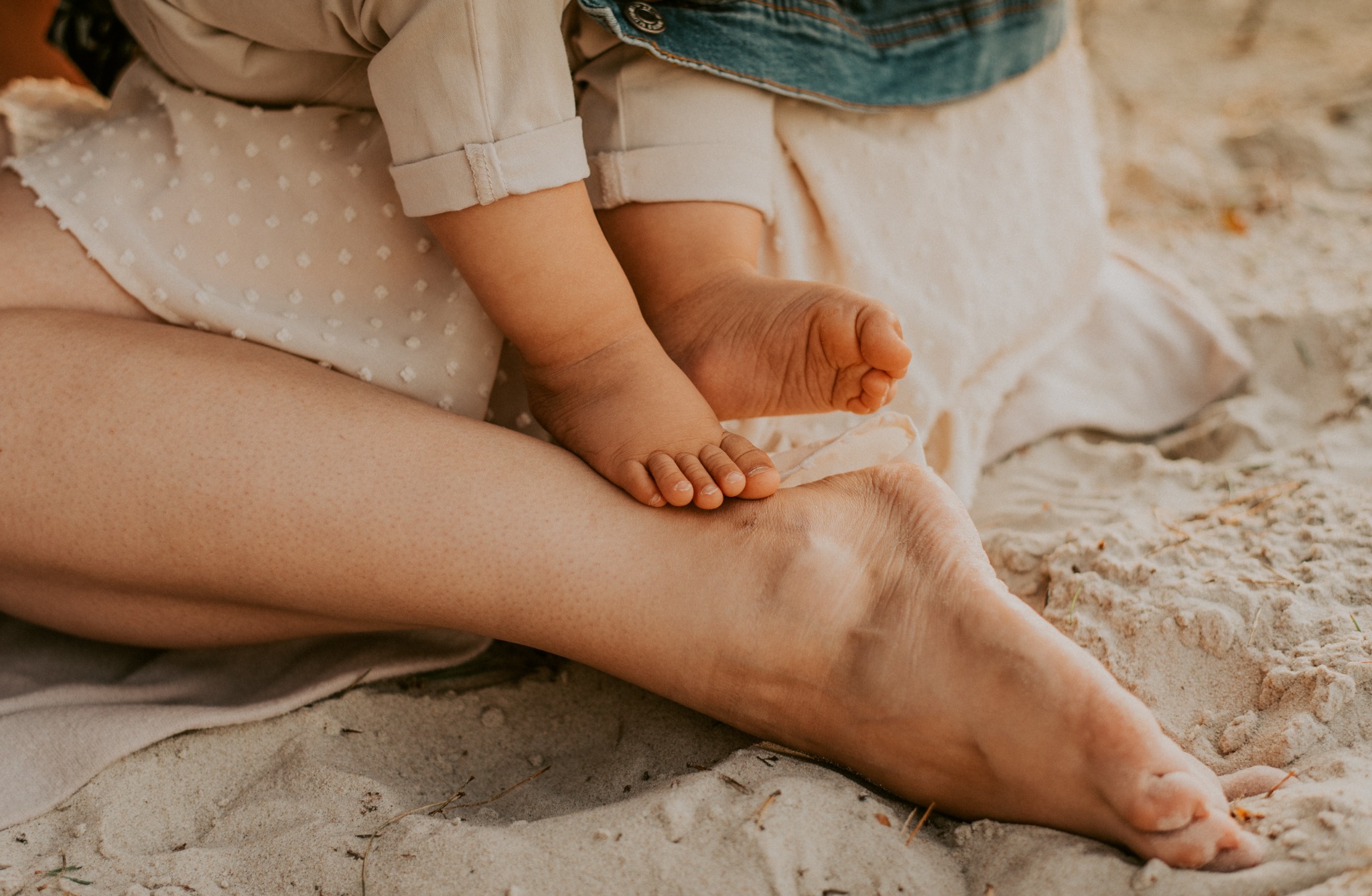 Drobne stópki dziecka spoczywają na stopie matki na tle piaszczystej plaży, delikatna sukienka w kropki i jeansowa kurtka dodają uroku scenie.