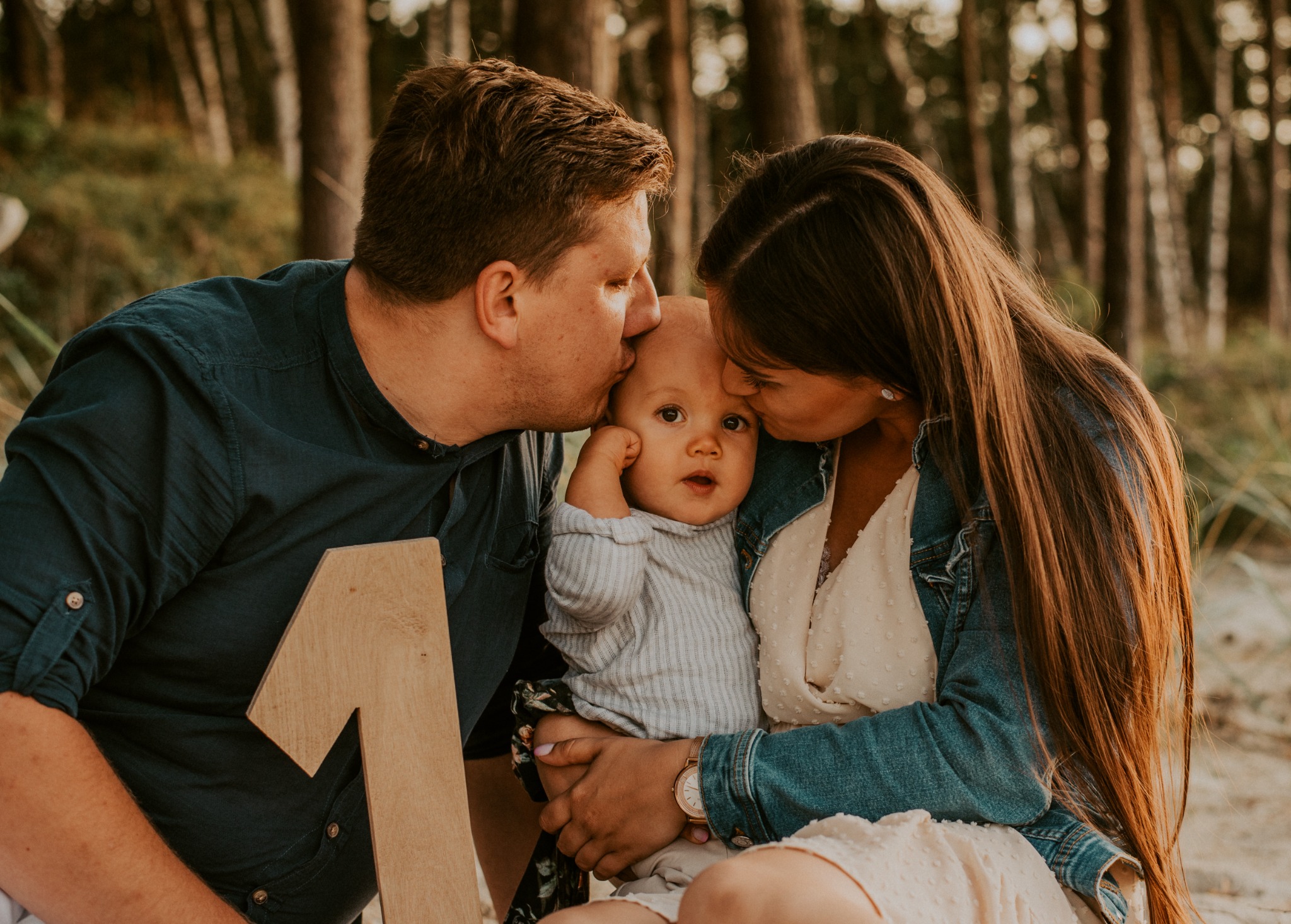 Rodzice całują rocznego synka w czoło na tle drzew; dziecko trzyma drewnianą cyfrę jeden.