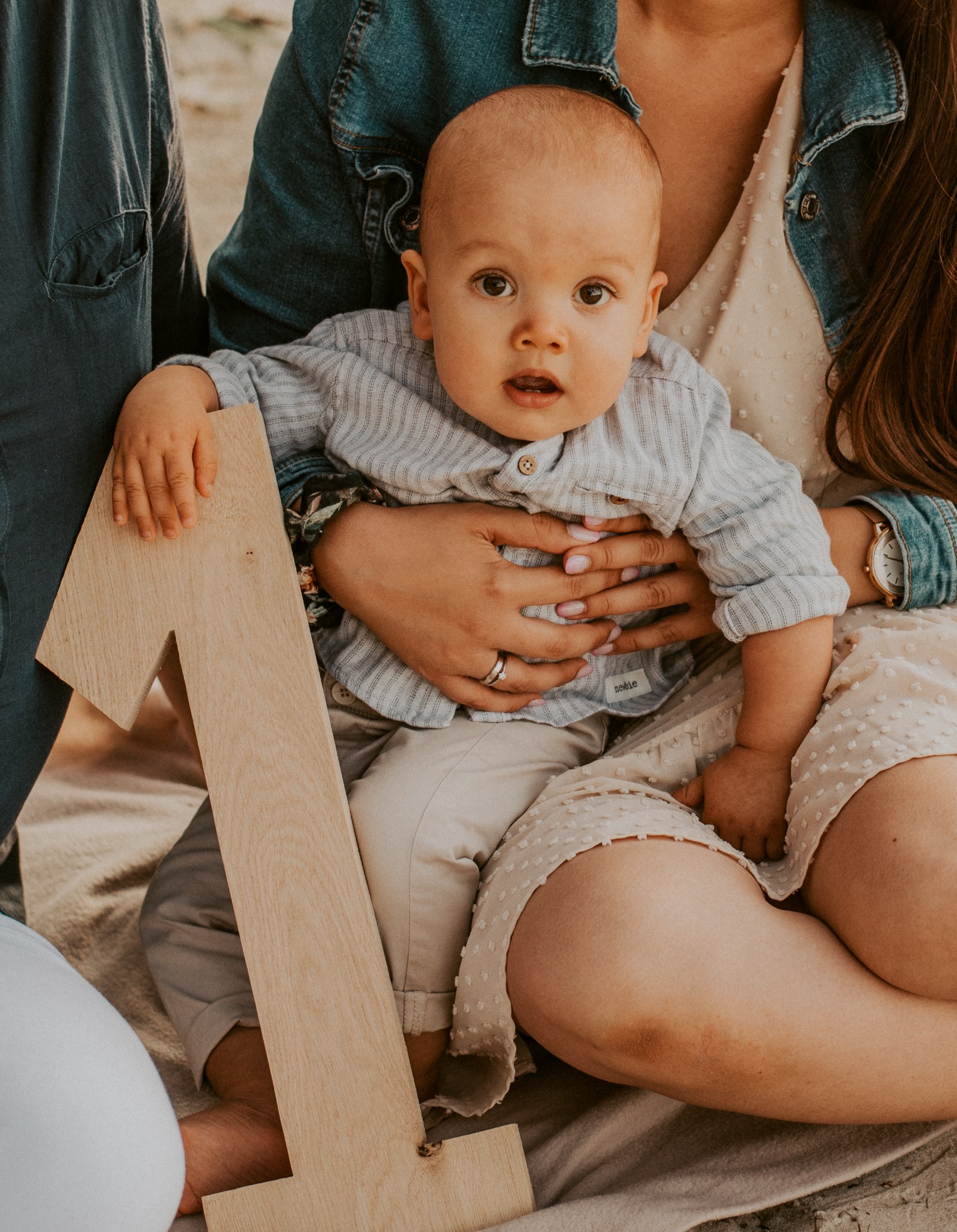 Ujęcie rocznego dziecka w ramionach matki, trzymającego drewnianą cyfrę jeden, w towarzystwie ojca, na tle piasku.