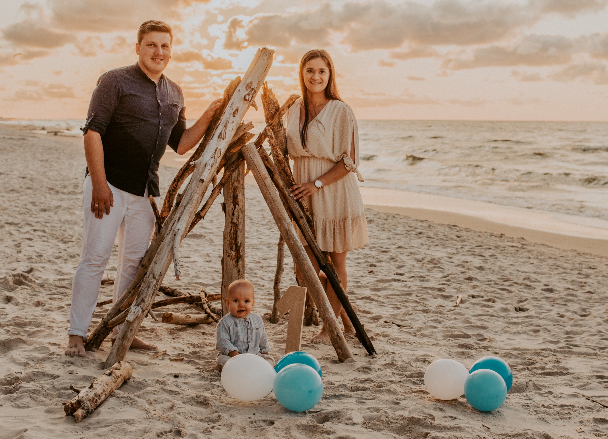 Rodzina pozuje na plaży przy konstrukcji z gałęzi i balonie z okazji pierwszych urodzin dziecka, w tle morze i zachodzące słońce.