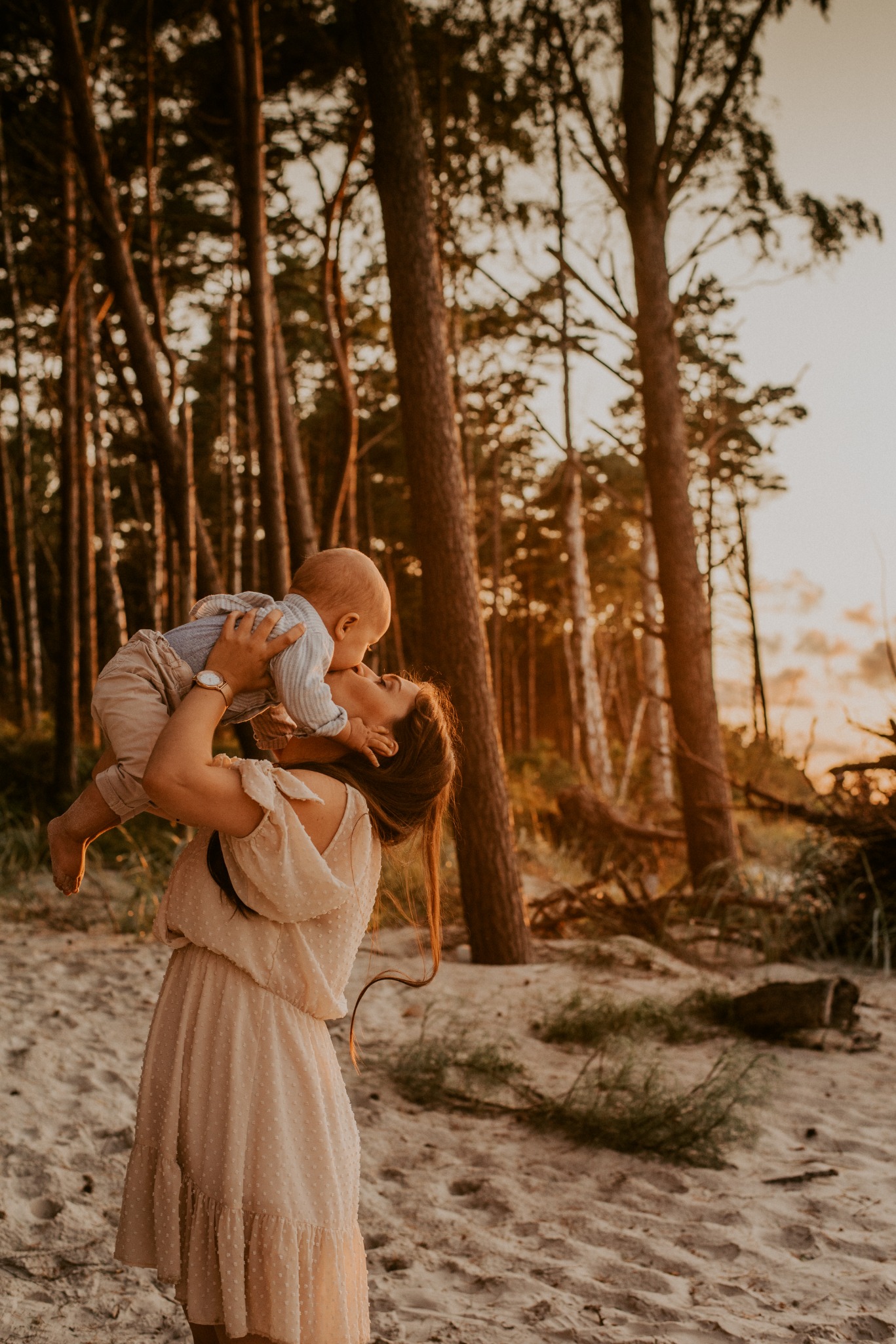 Matka w kremowej sukience unosi niemowlę w błękitnej koszuli i beżowych spodniach, całując je na tle piaszczystej plaży i lasu w ciepłym, zachodzącym słońcu.