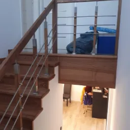 Drewniane schody z metalowymi barierkami w nowoczesnym domu, widok z dołu z widocznym fragmentem antresoli z workiem i pudłem.