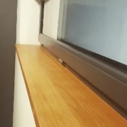 Jasny, drewniany parapet wewnętrzny pod oknem z szarą ramą, widoczny z boku.