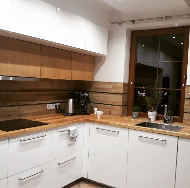 Nowoczesna kuchnia z białymi szafkami, drewnianym blatem i okładziną ścienną imitującą drewno, ekspres do kawy na blacie.