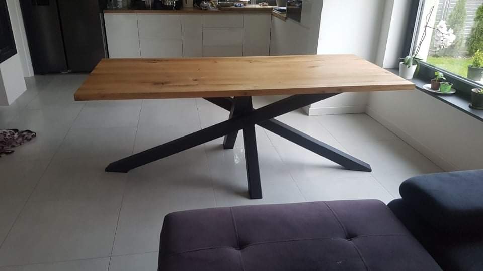 Stół z blatem z jasnego drewna i czarną, geometryczną podstawą w nowoczesnym wnętrzu z białymi szafkami kuchennymi i oknem z roślinami.