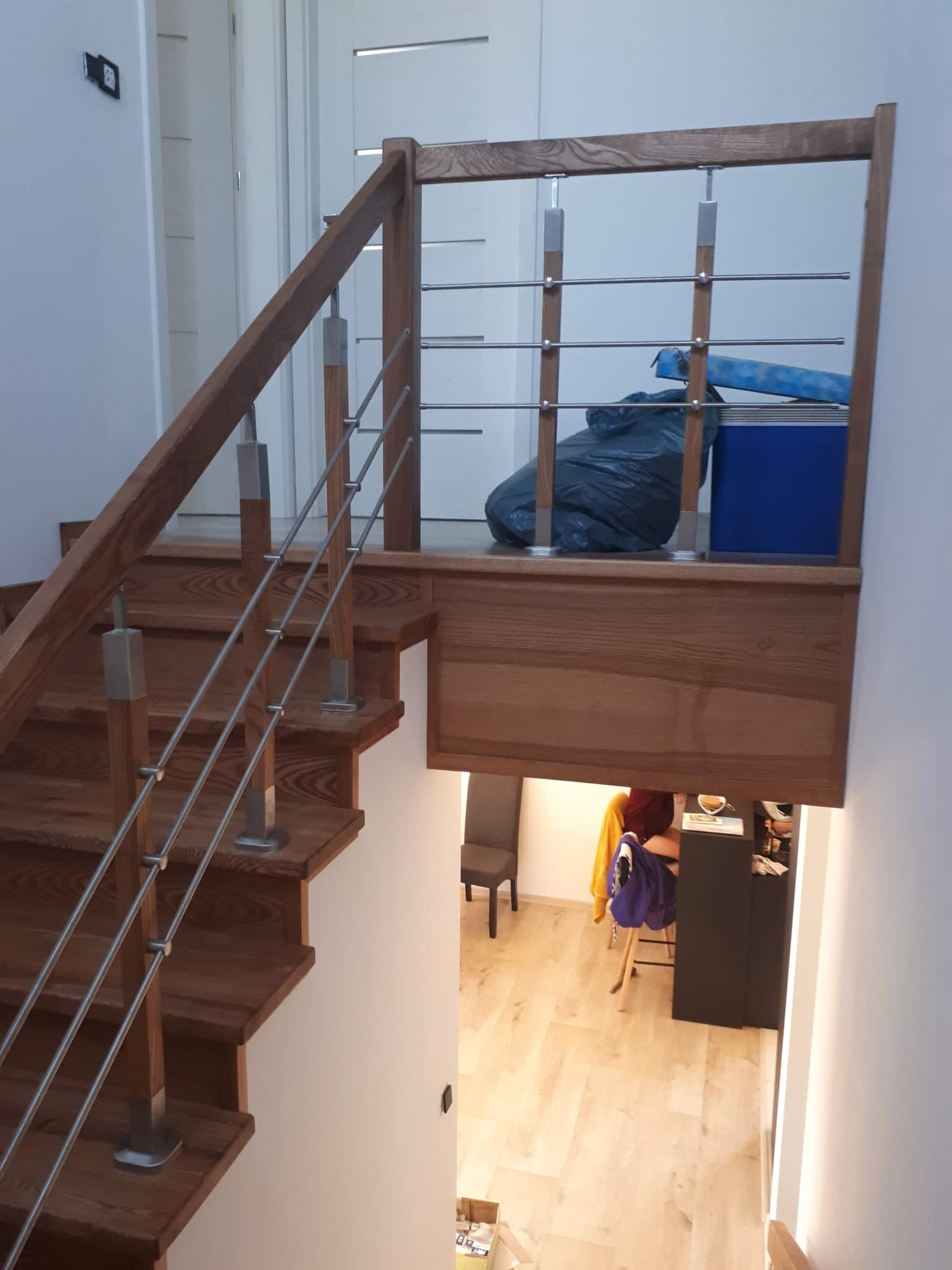 Drewniane schody z metalowymi barierkami w nowoczesnym domu, widok z dołu z widocznym fragmentem antresoli z workiem i pudłem.