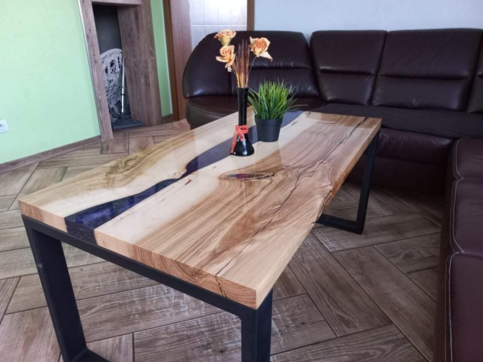 Stolik kawowy z blatem z jasnego drewna z żywicą epoksydową w kolorze ciemnego fioletu, na metalowych, czarnych nogach w kształcie prostokątów, ustawiony w salonie obok skórzanej kanapy.