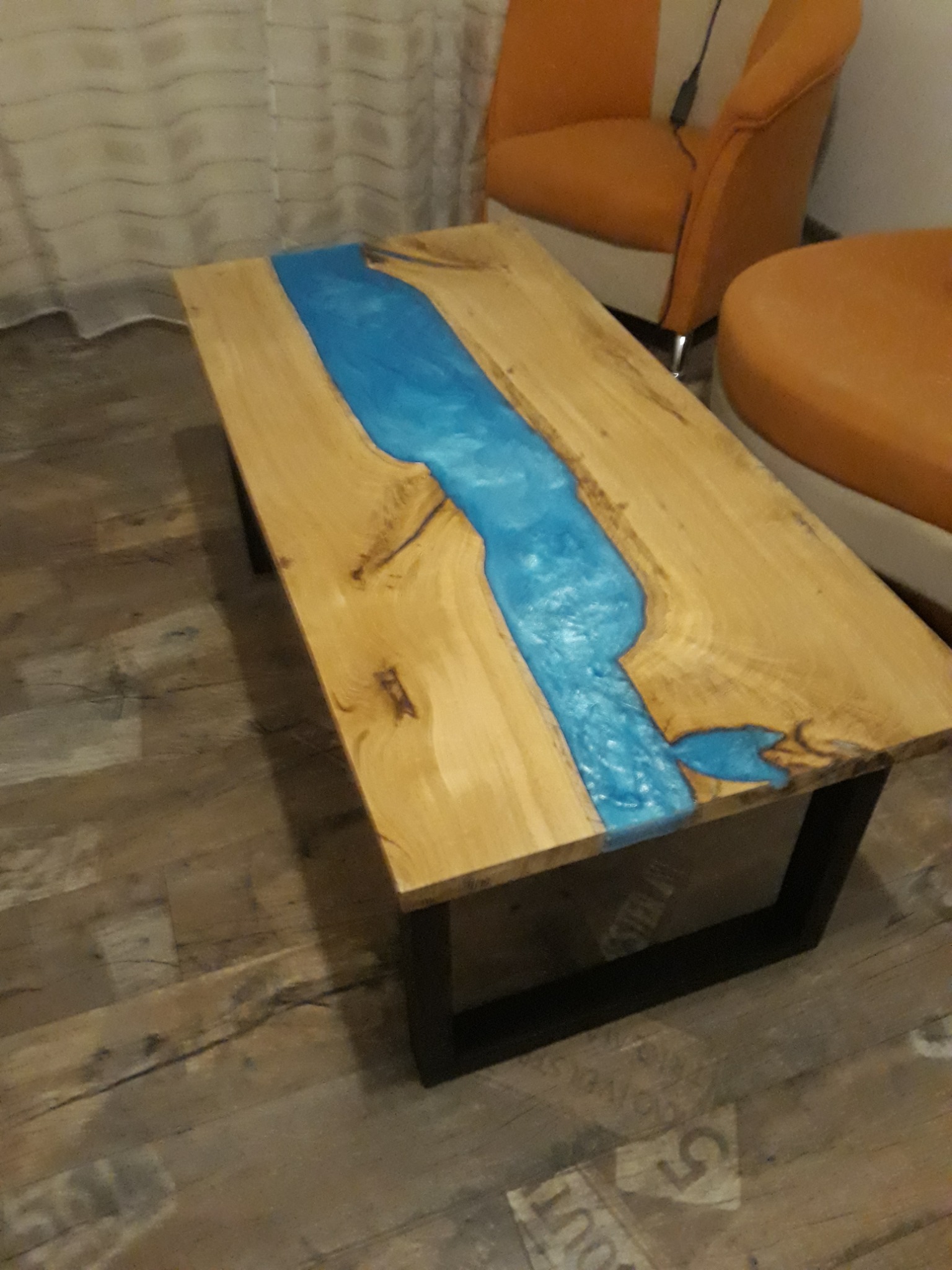 Stolik kawowy z blatem z jasnego drewna z żywicą epoksydową w kolorze błękitnym, imitującą rzekę, na czarnych, metalowych nogach, na drewnianej podłodze.