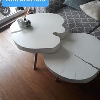 Biały, dwupoziomowy stolik kawowy o nieregularnym kształcie, przypominającym przekrój pnia drzewa, na drewnianej podłodze, z widocznym fragmentem sofy w tle.