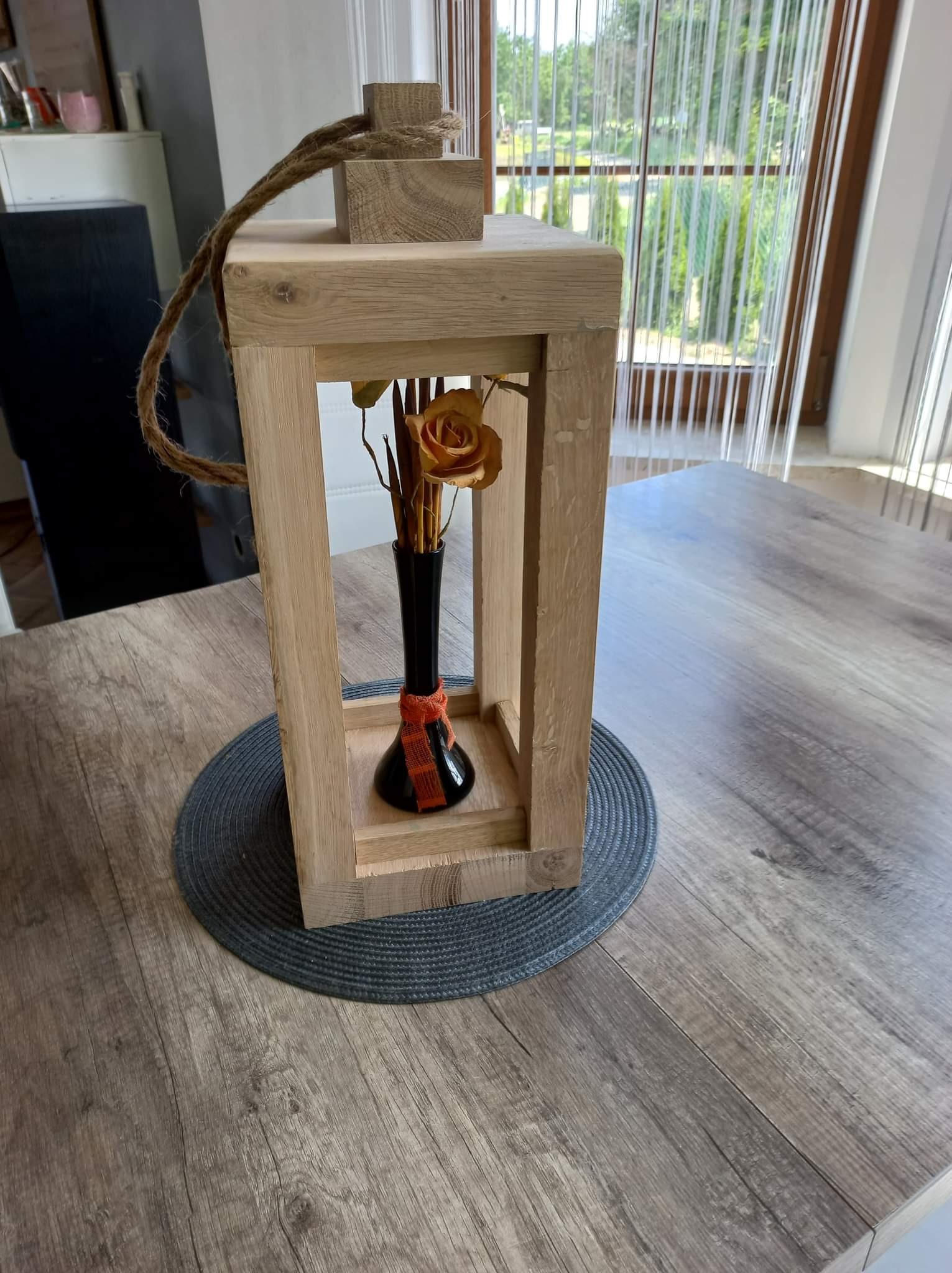 Dekoracyjny lampion z surowego drewna z jutowym sznurem, zawierający czarny wazon z pomarańczową różą, ustawiony na szarym podkładzie na drewnianym stole.