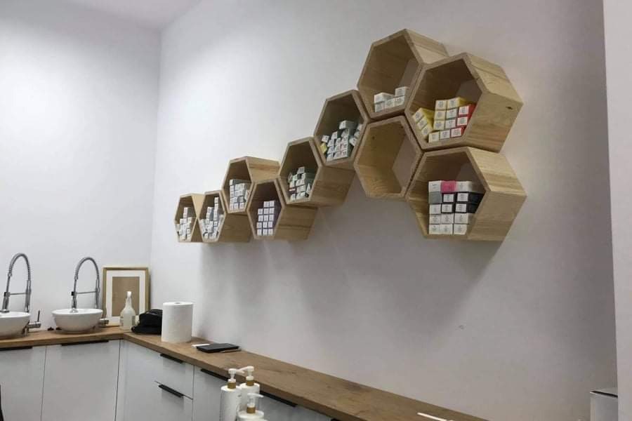 Ściana z drewnianymi, sześciokątnymi półkami w kształcie plastra miodu, wypełnionymi małymi pudełkami, nad blatem z umywalkami w jasnym pomieszczeniu.