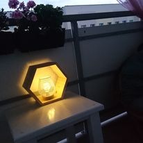 Sześciokątna drewniana lampka nocna z ciepłym, rozproszonym światłem, stojąca na białym stoliku na balkonie, w tle kwiaty w doniczce i wieczorne niebo.