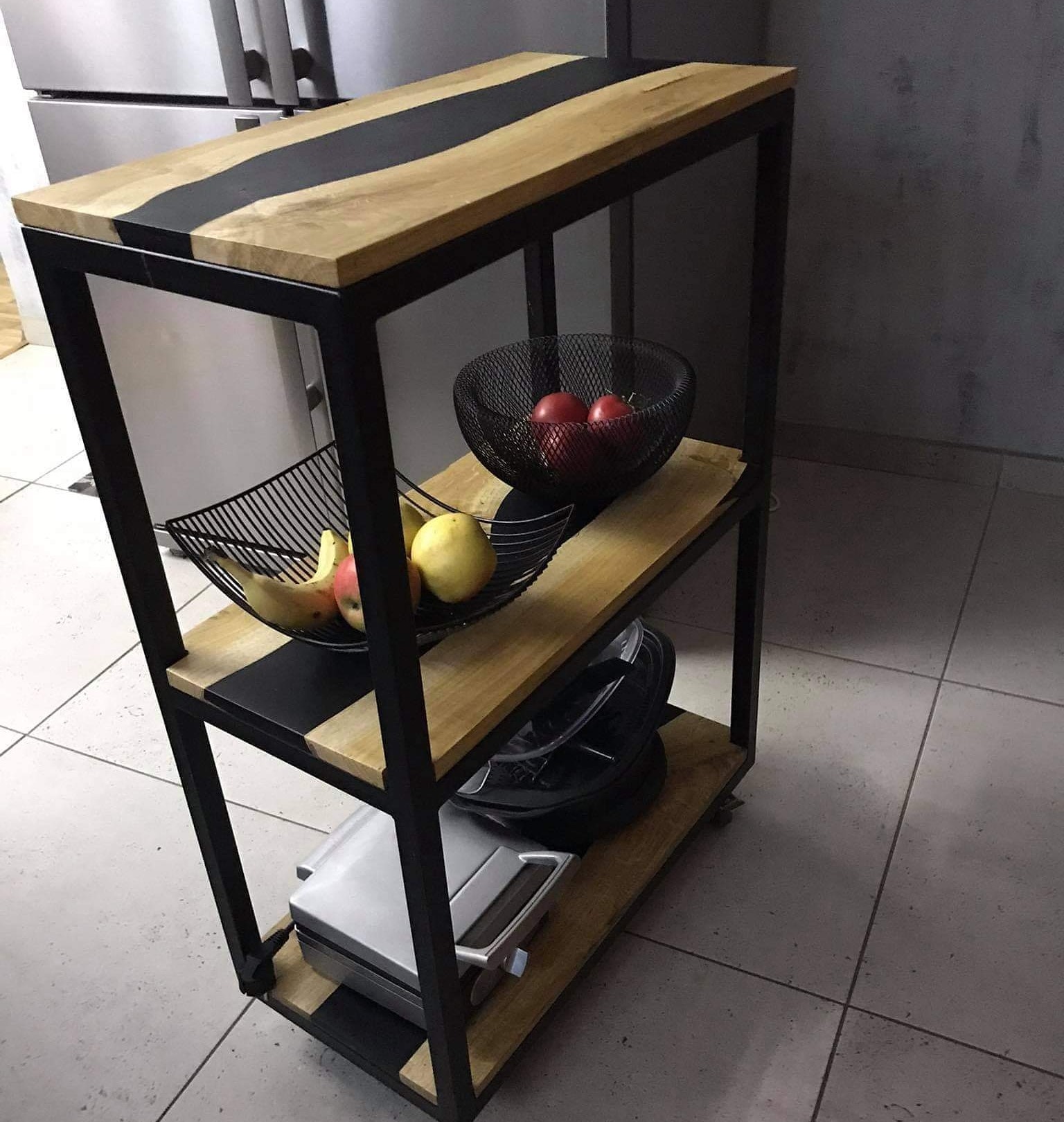 Wielopoziomowy regał kuchenny z drewnianymi półkami z czarnymi wstawkami, na kółkach, z owocami i urządzeniami kuchennymi na półkach, na tle lodówki i szarej ściany.