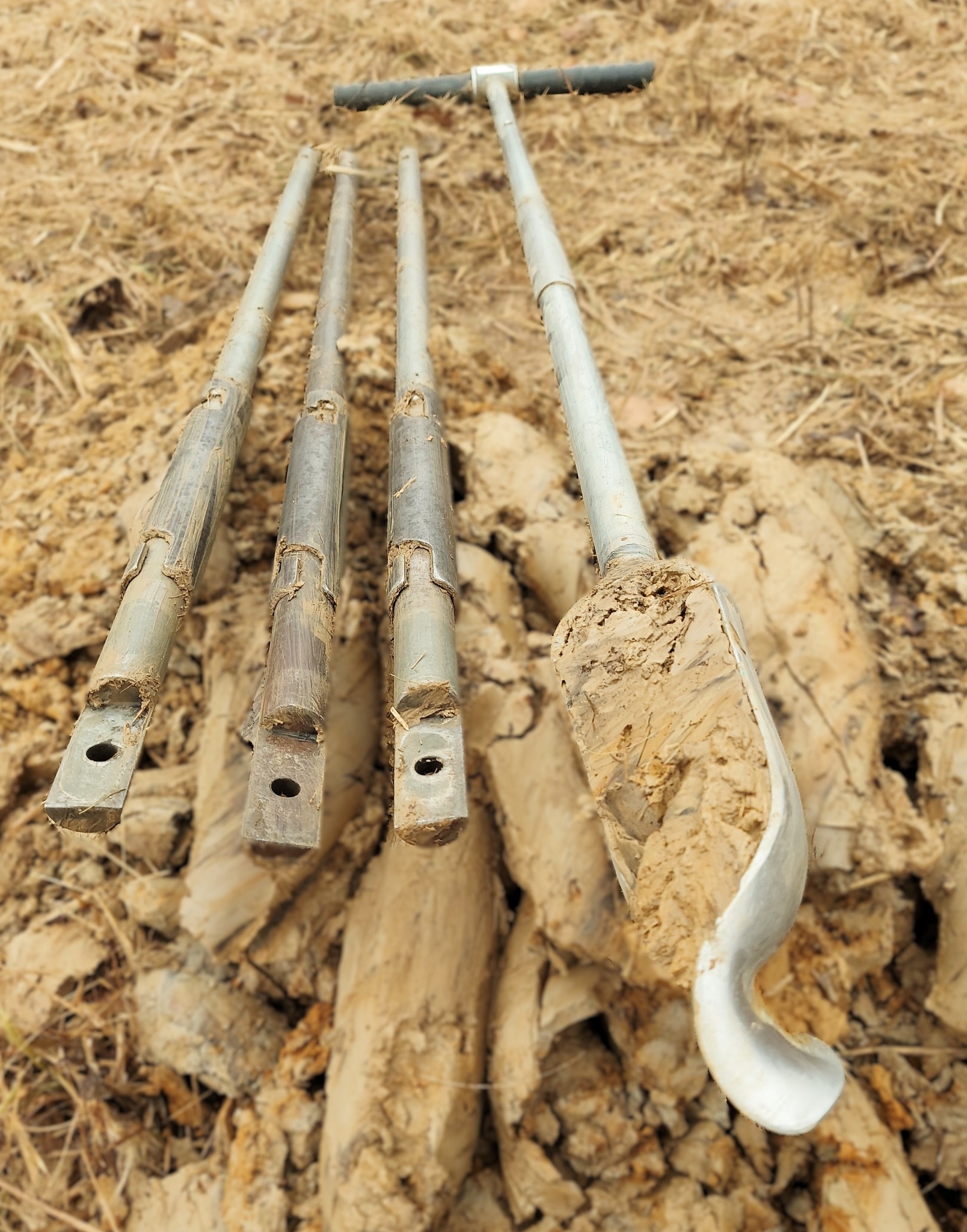 Wiertnica glebowa z próbkami ziemi. Metalowe rury z uchwytem T, widoczne ślady ziemi. Sprzęt do pobierania próbek gleby w terenie, analiza geologiczna.