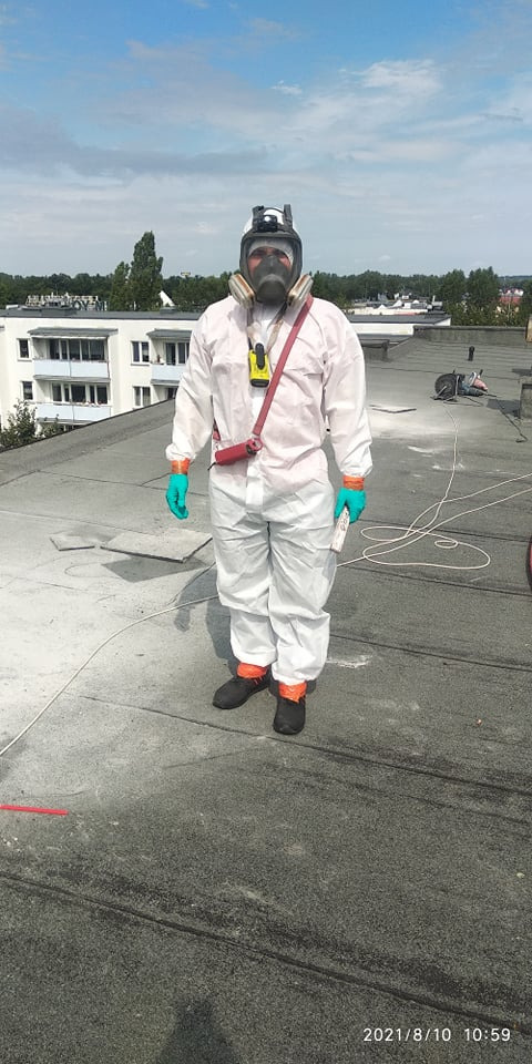 Pracownik w kombinezonie ochronnym i masce na dachu budynku, prawdopodobnie podczas inspekcji lub przygotowania do ocieplenia, z widocznym sprzętem pomiarowym i narzędziami w tle.