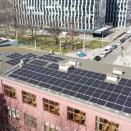 Widok z góry na dach budynku z zainstalowanymi panelami słonecznymi; w tle widoczne budynki miejskie i ulica z ruchem samochodowym, a na dachu robotnik w niebieskim kombinezonie.