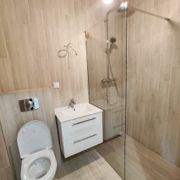 ProRemont - Nowoczesna łazienka z beżowymi płytkami, kabiną prysznicową typu walk-in, podwieszaną szafką z umywalką i toaletą z otwartą klapą.